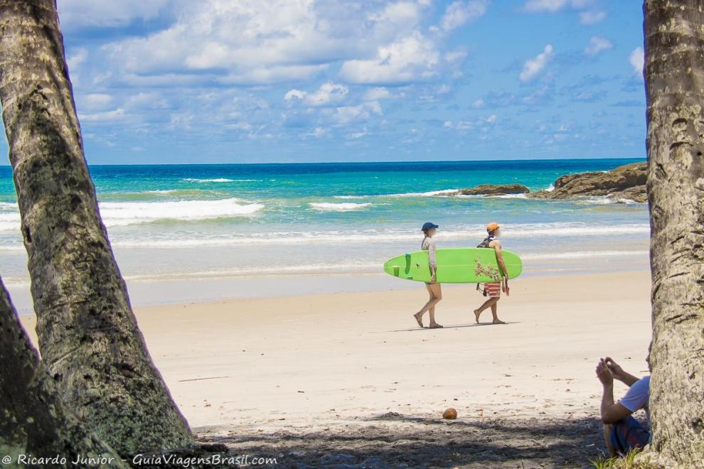 Imagem de duas pessoas levando prancha e o mar azulado na Praia da Engenhoca.